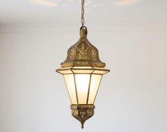 May include: Ornate brass pendant light with a geometric design. The lamp features a chain for hanging and a frosted glass shade. The intricate metalwork and warm glow create an inviting ambiance. A decorative lighting fixture for any room.