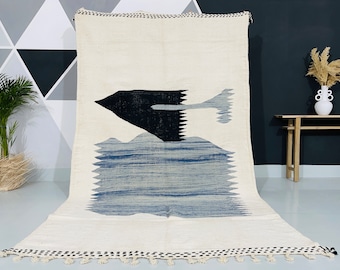May include: Cream-colored rug with a black and blue abstract design. The rug features a large black shape resembling a bird or plane, with a blue and gray reflection over blue horizontal lines. The edges have a black and white border.
