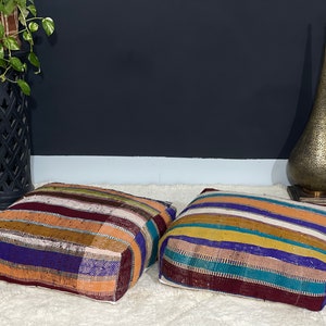 May include: Two colourful striped floor cushions with a woven pattern in shades of purple, orange, brown, white and teal. The cushions are sitting on a white fluffy rug.