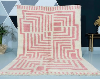 May include: A large, rectangular rug with a geometric pattern in pink and off-white. The design features a series of connected squares and rectangles. The rug is made of a soft, textured material, and is suitable for a living room or bedroom.