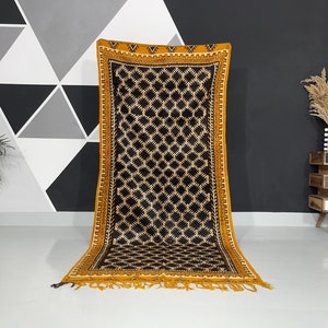 May include: A black and gold patterned rug with a geometric design. The rug has a fringe border and is laid out on a white floor.