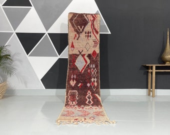May include: A rectangular, patterned rug with a beige, red, and brown color scheme. The rug features geometric diamond and zigzag designs. The rug is displayed vertically, showcasing its intricate details and texture. The rug has a fringed edge.