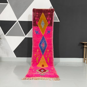 Può includere: Un vivace tappeto runner rosa con disegni geometrici blu, gialli e bordeaux. Il tappeto presenta un motivo a rombi e frange, che aggiungono un tocco bohémien. Il tappeto è esposto su un pavimento chiaro.