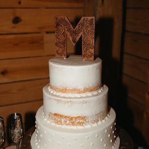 May include: A three-tiered white wedding cake with a wooden letter "M" topper decorated with intricate floral designs. The cake is on a silver cake stand with an ornate design.