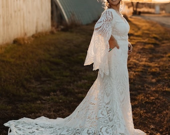 May include: A woman in a white lace wedding dress with a long train stands in a field with a wooden fence and a metal building in the background. The dress has a V-neckline and long sleeves with a bell shape. The woman is smiling and looking over her shoulder.