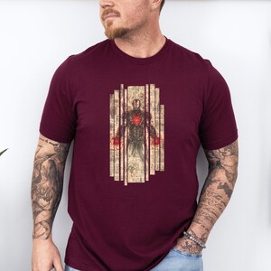 May include: Burgundy t-shirt featuring a detailed illustration of a superhero in a suit of armor. The graphic has a vintage, blueprint-like aesthetic with red accents. The design is centered on the chest of the shirt.