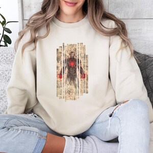 May include: Beige sweatshirt featuring a graphic of Iron Man. The design shows Iron Man in a standing pose, with a red and gold color scheme. The graphic is set against a background of vertical, textured panels.