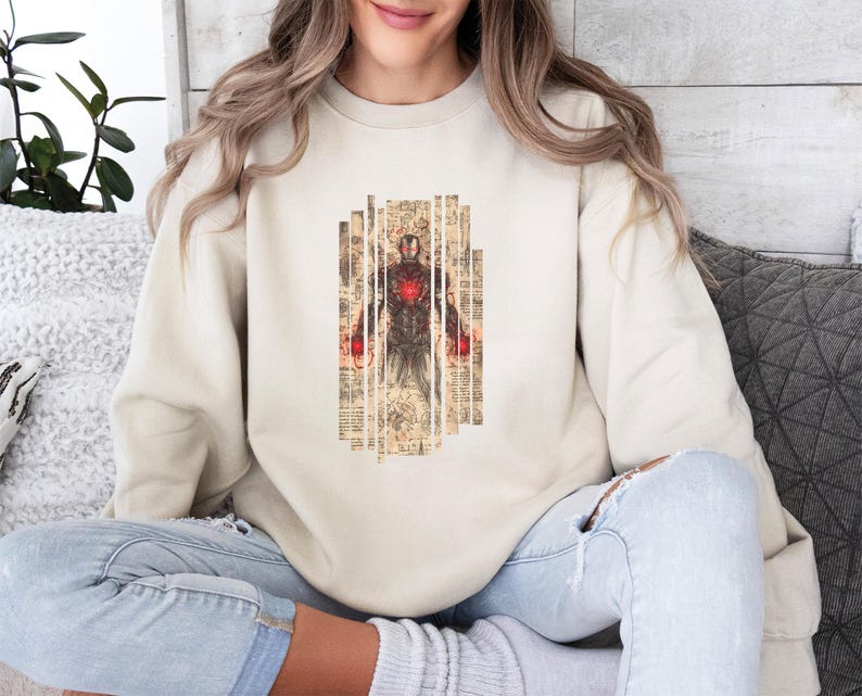 May include: Beige sweatshirt featuring a graphic of Iron Man. The design shows Iron Man in a standing pose, with a red and gold color scheme. The graphic is set against a background of vertical, textured panels.