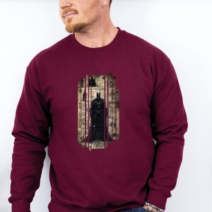 Puede incluir: Sudadera burdeos con un gr&aacute;fico de Batman. El gr&aacute;fico muestra a Batman de pie en un marco rectangular vertical con un fondo de paisaje urbano. La sudadera tiene mangas largas y cuello redondo.