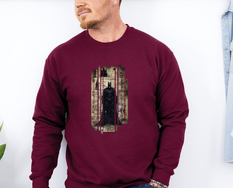 Puede incluir: Sudadera burdeos con un gr&aacute;fico de Batman. El gr&aacute;fico muestra a Batman de pie en un marco rectangular vertical con un fondo de paisaje urbano. La sudadera tiene mangas largas y cuello redondo.