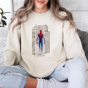Op de afbeelding: Een cr&egrave;mekleurige sweatshirt met een afbeelding van Spider-Man in een rood en blauw pak. De superheld wordt afgebeeld in verticale, rechthoekige panelen met een vintage papiertextuur. De sweatshirt is een casual, alledaags kledingstuk.