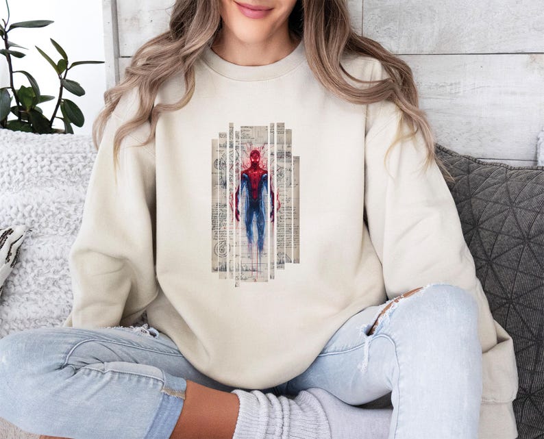 Op de afbeelding: Een cr&egrave;mekleurige sweatshirt met een afbeelding van Spider-Man in een rood en blauw pak. De superheld wordt afgebeeld in verticale, rechthoekige panelen met een vintage papiertextuur. De sweatshirt is een casual, alledaags kledingstuk.