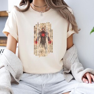 May include: Cream-colored t-shirt featuring a graphic of a figure with a red chest and hands, set against a backdrop of vintage paper. The design is centered on the shirt. The shirt has short sleeves.