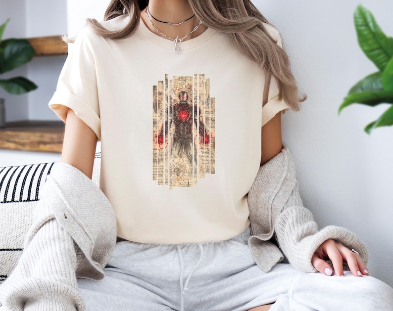 May include: Cream-colored t-shirt featuring a graphic of a figure with a red chest and hands, set against a backdrop of vintage paper. The design is centered on the shirt. The shirt has short sleeves.