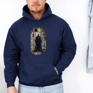 May include: Navy blue hooded sweatshirt featuring a Batman graphic. The graphic depicts Batman standing in a cage-like structure with a cityscape background. The sweatshirt has a front pocket and a drawstring hood.