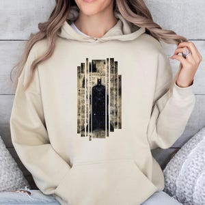 May include: A beige hoodie featuring a graphic of Batman. The design is a black silhouette of the character within a vertical rectangular frame. The frame has a vintage newspaper print background with white vertical lines.