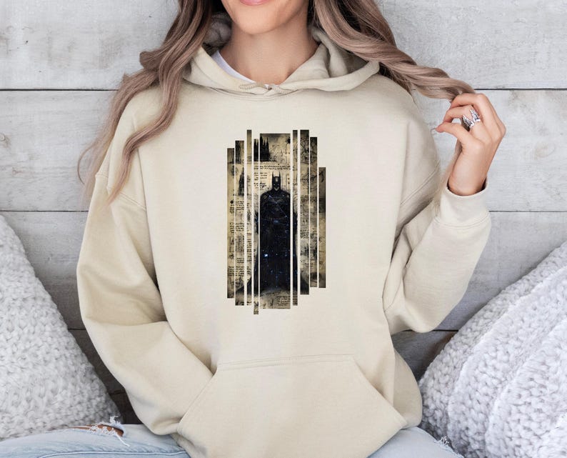 May include: A beige hoodie featuring a graphic of Batman. The design is a black silhouette of the character within a vertical rectangular frame. The frame has a vintage newspaper print background with white vertical lines.