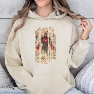 May include: Beige hoodie featuring a graphic of Iron Man in a vintage style. The design is centered on the front, showcasing the character in a detailed, artistic rendering. The hoodie has a drawstring hood and a kangaroo pocket.