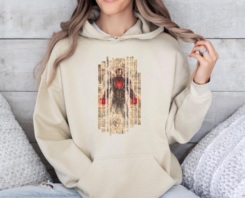 May include: Beige hoodie featuring a graphic of Iron Man in a vintage style. The design is centered on the front, showcasing the character in a detailed, artistic rendering. The hoodie has a drawstring hood and a kangaroo pocket.