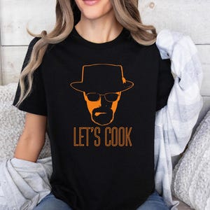 May include: Black t-shirt featuring an orange graphic of a man in a hat and glasses, with the words "LET'S COOK" in orange. The design is printed on the front. The shirt is made of a soft material.