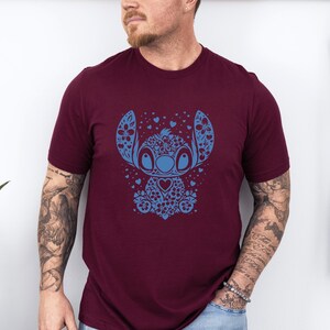 Puede incluir: Camiseta burdeos con un dise&ntilde;o floral azul de un personaje de dibujos animados. El personaje tiene ojos grandes, un pecho en forma de coraz&oacute;n y detalles florales. La camiseta es de manga corta y de material suave.