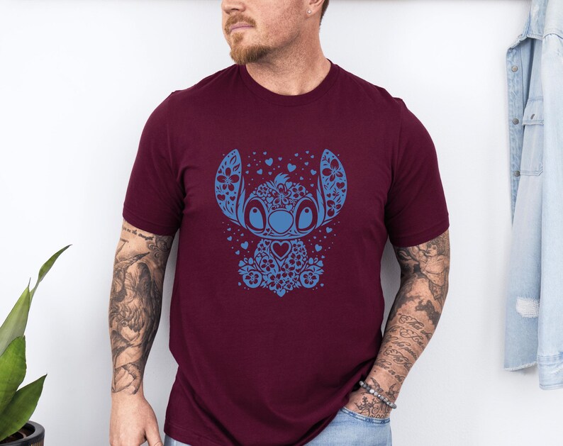 Puede incluir: Camiseta burdeos con un dise&ntilde;o floral azul de un personaje de dibujos animados. El personaje tiene ojos grandes, un pecho en forma de coraz&oacute;n y detalles florales. La camiseta es de manga corta y de material suave.