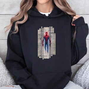 K&ouml;nnte beinhalten: Schwarzer Kapuzenpullover mit einem Spiderman-Motiv. Die Grafik zeigt Spiderman in einem rot-blauen Anzug, vor einem Hintergrund aus vertikalen Linien und Papier im Vintage-Stil. Der Kapuzenpullover hat eine Kordelkapuze und lange &Auml;rmel.