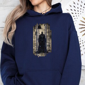 May include: Navy blue hoodie featuring a Batman graphic. The graphic depicts Batman standing behind bars, with a vintage-style background. The hoodie has a front pocket and a drawstring hood.