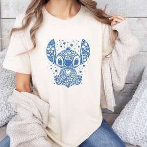 Puede incluir: Camiseta color crema con un dise&ntilde;o floral azul de un personaje de dibujos animados. El dise&ntilde;o incluye corazones y patrones florales. La camiseta tiene mangas cortas y cuello redondo.