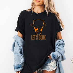 May include: Black t-shirt with an orange graphic of a man in a hat and glasses, and the orange text "LET'S COOK". The t-shirt is a relaxed fit, ideal for casual wear.