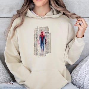 K&ouml;nnte beinhalten: Beiger Kapuzenpullover mit einer Grafik von Spider-Man in Rot und Blau. Der Superheld ist in einem vertikalen rechteckigen Design mit einem strukturierten, Vintage-Hintergrund dargestellt. Der Hoodie hat eine Vordertasche und eine Kapuze mit Kordelzug.