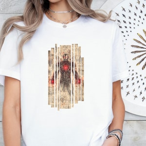 May include: White t-shirt featuring a graphic of a superhero in a suit, with a red chest piece and red accents on the hands. The graphic is set against a backdrop of vertical, vintage-style paper strips. The design has a vintage, comic book feel.