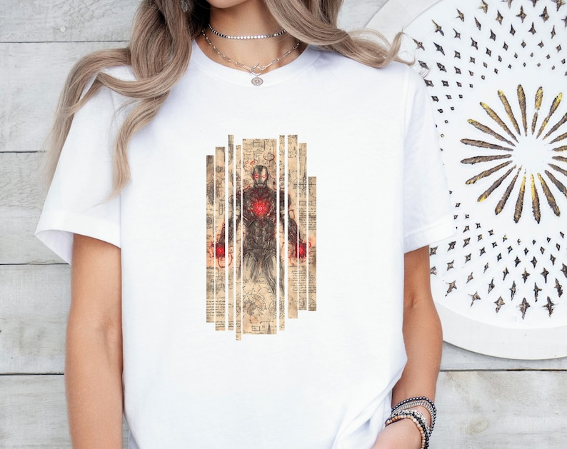 May include: White t-shirt featuring a graphic of a superhero in a suit, with a red chest piece and red accents on the hands. The graphic is set against a backdrop of vertical, vintage-style paper strips. The design has a vintage, comic book feel.