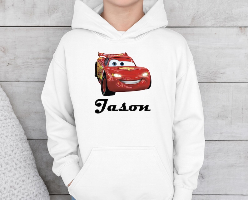 Puede incluir: Sudadera con capucha blanca con un coche de carreras rojo de dibujos animados con una cara sonriente. El nombre "Jason" est&aacute; impreso en negro debajo. La sudadera tiene un bolsillo delantero y una capucha con cord&oacute;n.