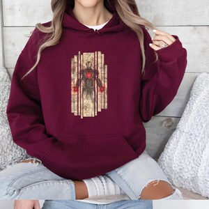 May include: Burgundy hoodie featuring a graphic of a superhero in a suit with red accents. The superhero is set against a backdrop of vertical lines and vintage-style paper. The hoodie has a drawstring hood and a kangaroo pocket.