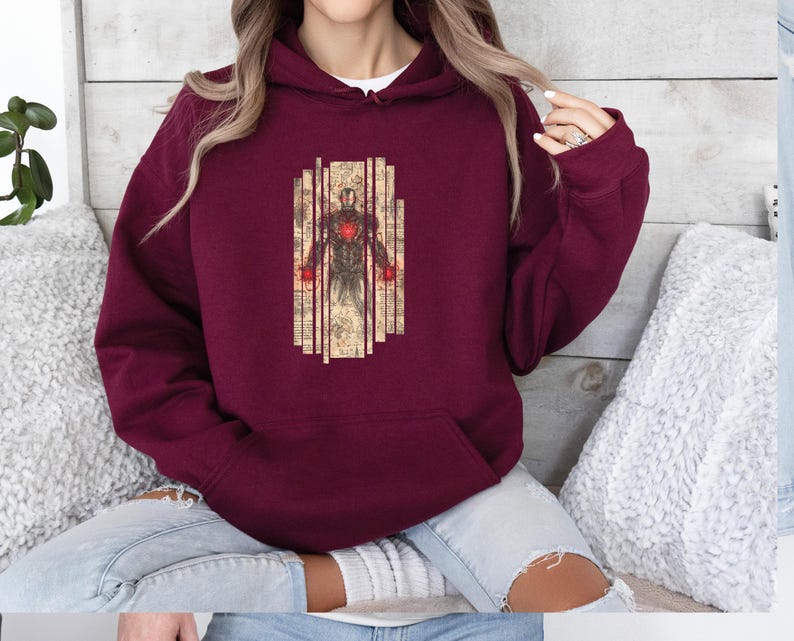 May include: Burgundy hoodie featuring a graphic of a superhero in a suit with red accents. The superhero is set against a backdrop of vertical lines and vintage-style paper. The hoodie has a drawstring hood and a kangaroo pocket.