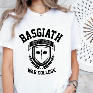 May include: White t-shirt with black text that reads "Basgiath Est 2023 War College". The text is in a stylized font and is surrounded by a crest with a dragon and laurel leaves.