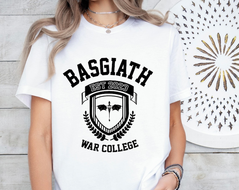 May include: White t-shirt with black text that reads "Basgiath Est 2023 War College". The text is in a stylized font and is surrounded by a crest with a dragon and laurel leaves.