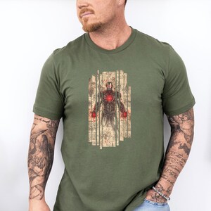 May include: Olive green t-shirt featuring a detailed illustration of Iron Man. The graphic shows Iron Man's armor with a red chest piece and red hands, set against a backdrop of aged paper. The shirt is a crew neck.