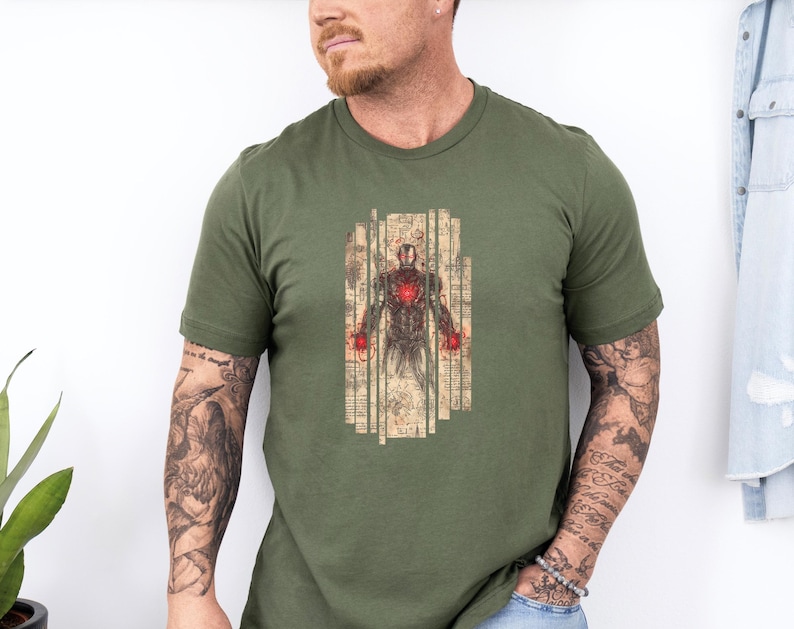 May include: Olive green t-shirt featuring a detailed illustration of Iron Man. The graphic shows Iron Man's armor with a red chest piece and red hands, set against a backdrop of aged paper. The shirt is a crew neck.