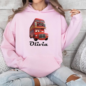 Puede incluir: Una sudadera con capucha rosa claro con un dibujo animado de un autob&uacute;s de dos pisos rojo, con el nombre "Olivia" en escritura negra. El autob&uacute;s tiene una cara sonriente y detalles amarillos. La sudadera tiene un bolsillo delantero y una capucha con cord&oacute;n.