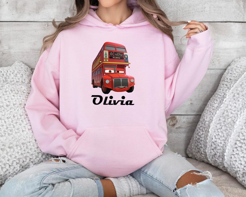 Puede incluir: Una sudadera con capucha rosa claro con un dibujo animado de un autob&uacute;s de dos pisos rojo, con el nombre "Olivia" en escritura negra. El autob&uacute;s tiene una cara sonriente y detalles amarillos. La sudadera tiene un bolsillo delantero y una capucha con cord&oacute;n.