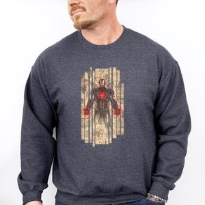 May include: A dark gray sweatshirt featuring a detailed illustration of Iron Man. The graphic is in a vertical rectangular shape with a vintage, aged paper look. The Iron Man design has red and gold accents.