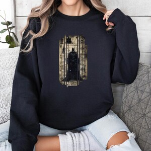 Puede incluir: Sudadera negra con un gr&aacute;fico de Batman. El gr&aacute;fico muestra a Batman dentro de un marco vertical, sobre un fondo de papel vintage. La sudadera es de color negro liso y tiene mangas largas.
