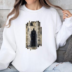 Puede incluir: Sudadera blanca con un gr&aacute;fico de Batman. El gr&aacute;fico es una silueta negra de Batman sobre un fondo de papel vintage y rayas verticales. Una prenda casual y c&oacute;moda.