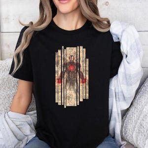 May include: Black t-shirt featuring a detailed illustration of Iron Man. The design includes a central figure of Iron Man with a glowing red chest and hands, set against a backdrop of vertical, aged paper-like panels. The artwork has a vintage, sketched aesthetic.