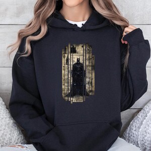 May include: Black hooded sweatshirt featuring a Batman graphic. The graphic is a stylized image of Batman within a rectangular frame with a vintage, aged paper background. The design is in shades of black, gray, and beige.