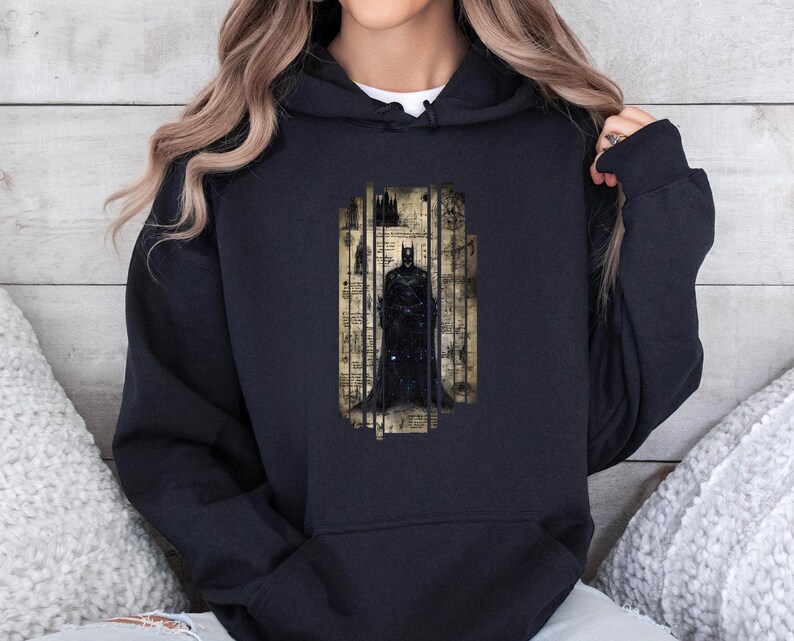 May include: Black hooded sweatshirt featuring a Batman graphic. The graphic is a stylized image of Batman within a rectangular frame with a vintage, aged paper background. The design is in shades of black, gray, and beige.
