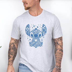 Puede incluir: Camiseta gris claro con un dise&ntilde;o floral azul de un personaje de dibujos animados. El dise&ntilde;o incluye elementos florales, corazones y un centro en forma de coraz&oacute;n. La camiseta es de cuello redondo.