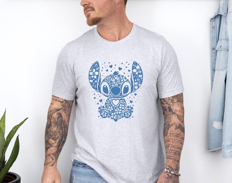 Puede incluir: Camiseta gris claro con un dise&ntilde;o floral azul de un personaje de dibujos animados. El dise&ntilde;o incluye elementos florales, corazones y un centro en forma de coraz&oacute;n. La camiseta es de cuello redondo.